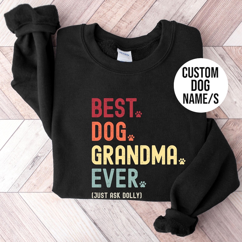 Puede incluir: Sudadera negra con el texto "BEST DOG GRANDMA EVER" en rojo, naranja, amarillo y azul. El texto incluye huellas de patas y la frase "(JUST ASK DOLLY)". Un c&iacute;rculo blanco dice "CUSTOM DOG NAME/S".