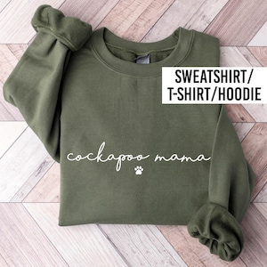 May include: An olive green long-sleeve sweatshirt featuring the white text "cockapoo mama" in a cursive script font, accompanied by a small white paw print icon below the text. The garment includes a crew neckline and ribbed cuffs.