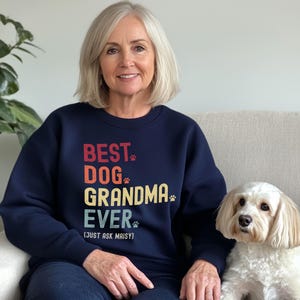 Puede incluir: Una sudadera azul marino con el texto "BEST DOG GRANDMA EVER (JUST ASK MAISY)" en rojo, naranja, amarillo y verde. La sudadera tiene huellas de patas incorporadas en el dise&ntilde;o. Un perro blanco est&aacute; sentado al lado de la persona que lleva la sudadera.