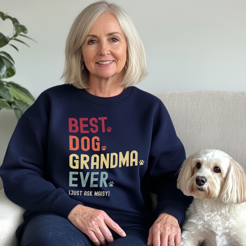 Puede incluir: Una sudadera azul marino con el texto "BEST DOG GRANDMA EVER (JUST ASK MAISY)" en rojo, naranja, amarillo y verde. La sudadera tiene huellas de patas incorporadas en el dise&ntilde;o. Un perro blanco est&aacute; sentado al lado de la persona que lleva la sudadera.