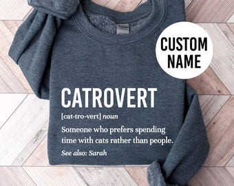 May include: A dark gray sweatshirt with the text "CATROVERT [cat-tro-vert] noun Someone who prefers spending time with cats rather than people. See also: Sarah" printed on the front. The text is in white. The sweatshirt is folded and lying on a wooden surface.