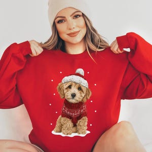 May include: A red sweatshirt featuring a cute puppy wearing a Santa hat and a red sweater with a white snowflake design. White dots are scattered around the puppy. The sweatshirt is a solid red color.