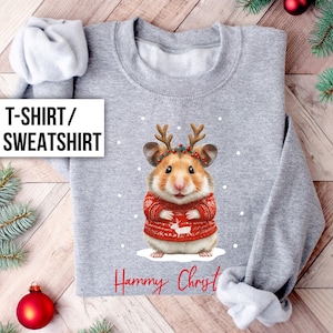 May include: A light grey sweatshirt with a festive hamster wearing a red sweater and antlers. The sweater has a reindeer design. The text "Hammy Christ" is printed in red below the hamster. The words "T-SHIRT / SWEATSHIRT" are in a white box.