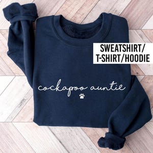 May include: A navy blue long-sleeve sweatshirt featuring the white text "cockapoo auntie" written in a cursive script font, accompanied by a small white paw print icon. A white text box in the upper right corner displays "SWEATSHIRT/T-SHIRT/HOODIE".