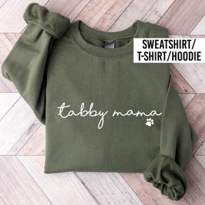 May include: An olive green sweatshirt with the words "tabby mama" in white script, accompanied by a small paw print. The image also includes the text "SWEATSHIRT/ T-SHIRT/HOODIE" in a white rectangle.