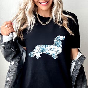 May include: A black short-sleeved t-shirt featuring a graphic of a long-haired dachshund silhouette filled with a blue and white floral pattern. The shirt is worn under a dark gray denim jacket with silver buttons and a gold chain necklace.