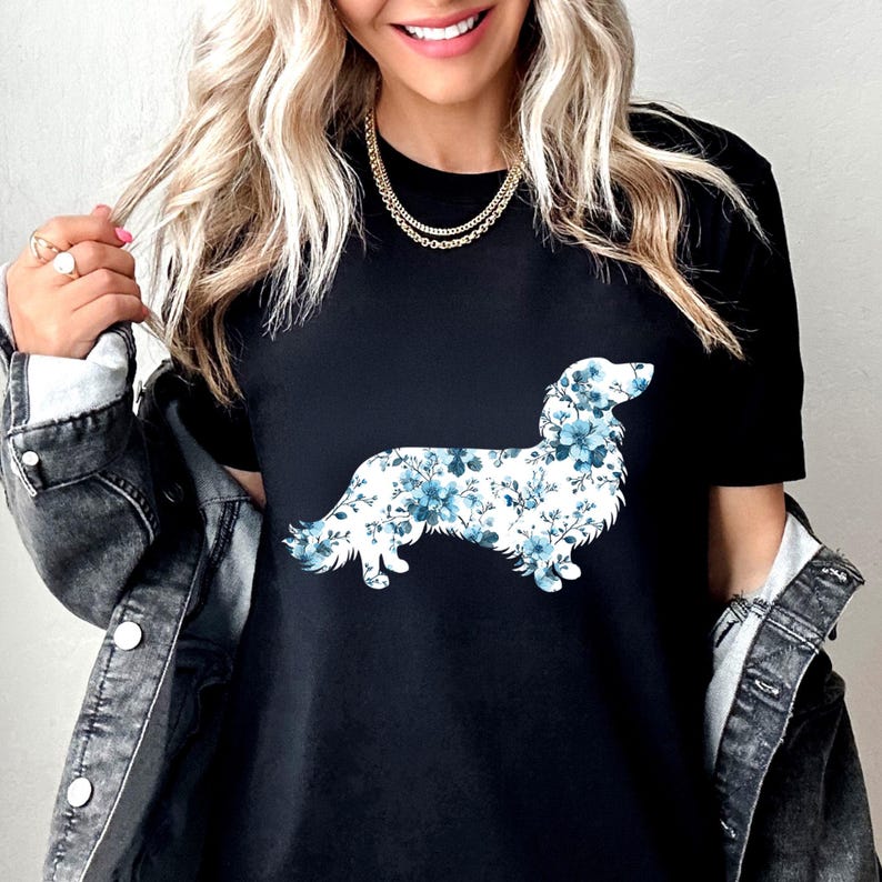 May include: A black short-sleeved t-shirt featuring a graphic of a long-haired dachshund silhouette filled with a blue and white floral pattern. The shirt is worn under a dark gray denim jacket with silver buttons and a gold chain necklace.