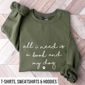 May include: An olive green crewneck sweatshirt featuring white cursive text that reads all i need is a book and my dog, accompanied by a small paw print icon centered below the text. The garment has long sleeves with ribbed cuffs and a ribbed neckline.