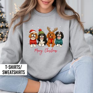 May include: A light gray sweatshirt featuring a graphic of four Cavalier King Charles Spaniels wearing Christmas sweaters and hats. The text "Merry Christmas" is written in red script below the dogs. The bottom left corner has the text "T-SHIRTS/ SWEATSHIRTS".
