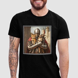 May include: A black t-shirt featuring a square graphic print of a medieval knight in silver armor and a red cape, holding four cats of various colors. The background of the graphic depicts a stone castle with towers under a cloudy sky.