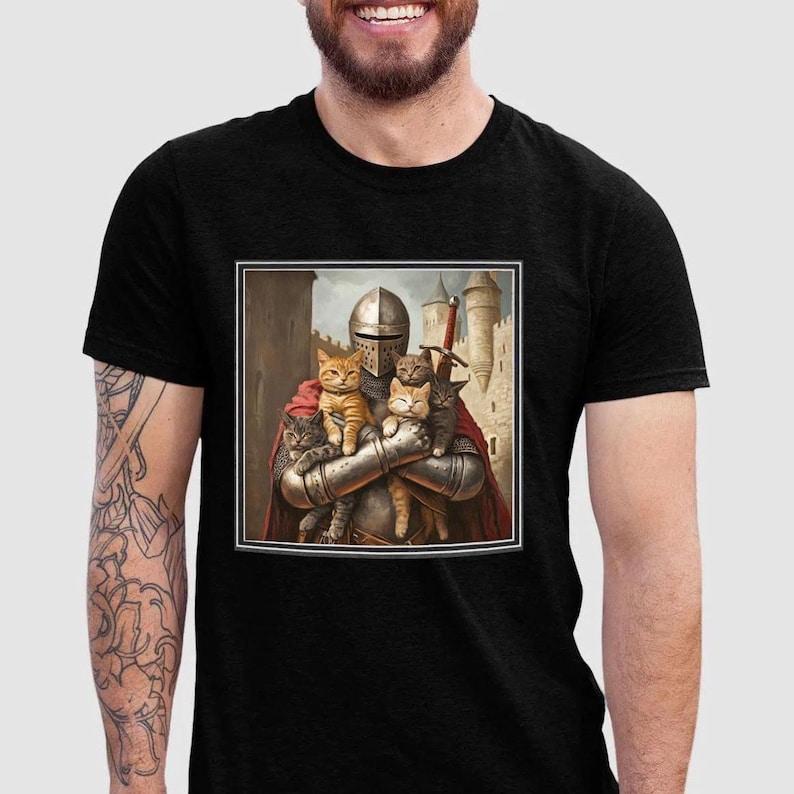 May include: A black t-shirt featuring a square graphic print of a medieval knight in silver armor and a red cape, holding four cats of various colors. The background of the graphic depicts a stone castle with towers under a cloudy sky.