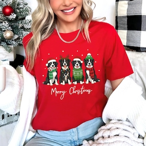 May include: A red t-shirt featuring four Border Collie dogs dressed in Christmas attire. Each dog wears a Santa hat, scarf, or sweater. The words "Merry Christmas" are written in white script below the dogs.