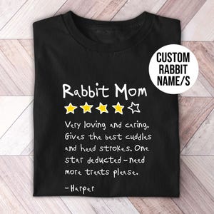 May include: A black t-shirt featuring the white text Rabbit Mom, four yellow stars, and one white star outline. Below, white text reads: Very loving and caring. Gives the best cuddles and head strokes. One star deducted - need more treats please. - Harper.