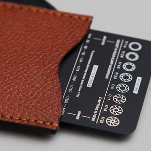 May include: A brown leather card holder with a black card partially inserted. The card has white text and aperture symbols. The leather has a textured surface and is stitched along the edges. The card displays aperture and shutter speed information.