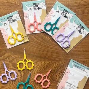 May include: A selection of embroidery scissors in shades of yellow, pink, teal, and purple. Each pair is packaged with a card that reads "PATTERN PAPER SCISSORS". The scissors have decorative handles and sharp blades. Made in the UK.