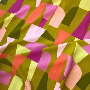 May include: Close-up of a fabric with a geometric pattern. The design features abstract shapes in shades of olive green, pink, orange, and white. The fabric has a soft, flowing appearance, with visible folds and creases.