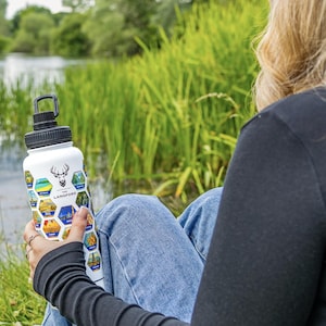 May include: White water bottle with a black cap and handle. The bottle features a deer logo and the text "LANGFORD" with colourful hexagonal designs. The bottle is held outdoors near a body of water and tall green grass.