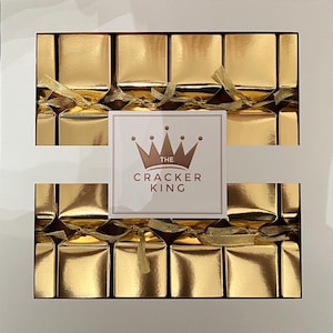 Puede incluir: Una caja de 18 crackers navideños dorados con una cinta dorada y un logotipo de corona dorada que dice "The Cracker King".