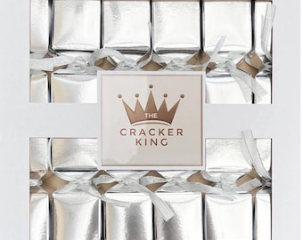 May include: A white box containing eight silver metallic party crackers arranged in two rows. Each cracker is tied with a silver ribbon. A square label in the center features a brown crown icon and the text THE CRACKER KING in brown lettering.