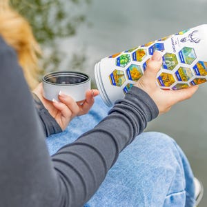 Op de afbeelding: Witte reismok met een zwart interieur en een deksel. De mok is versierd met kleurrijke zeshoekige afbeeldingen van bezienswaardigheden en de tekst "THE LANGFORD" en een hertenlogo. De mok wordt vastgehouden in de buurt van een watermassa.