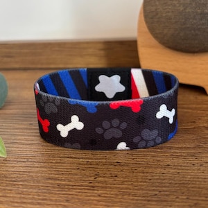 May include: A black dog collar featuring white bones, paw prints, red accents, and blue stripes. The collar sits on a wooden surface, alongside a wooden elephant figurine and a green plant.