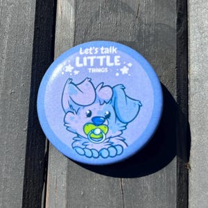 May include: A round, blue button with the text "Let's talk LITTLE THINGS" and a cartoon dog character with a pacifier. The button is on a weathered wooden surface.