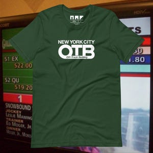 May include: A dark green t-shirt featuring the white text "NEW YORK CITY OTB Off-Track Betting". The shirt has a crew neck and short sleeves. The background shows a screen with race information.