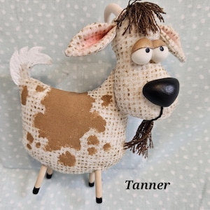 May include: A whimsical stuffed goat with a tan and white patterned body, brown spots, and a black nose. The goat has wooden legs and a small brown beard. The name "Tanner" is printed on the bottom right.