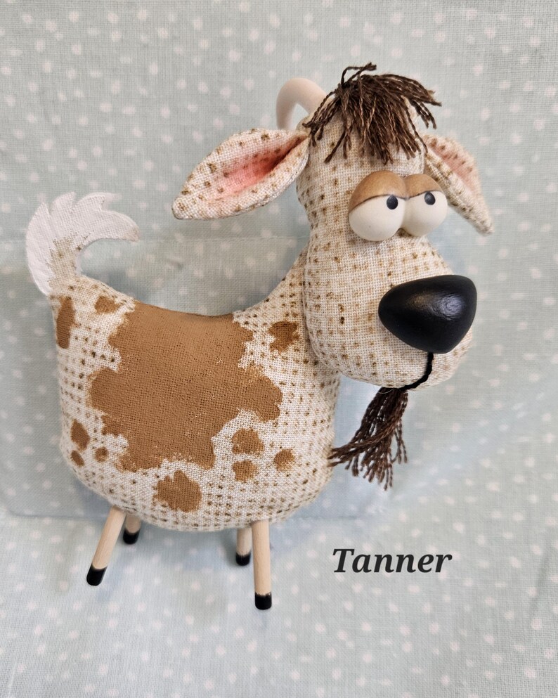 May include: A whimsical stuffed goat with a tan and white patterned body, brown spots, and a black nose. The goat has wooden legs and a small brown beard. The name "Tanner" is printed on the bottom right.