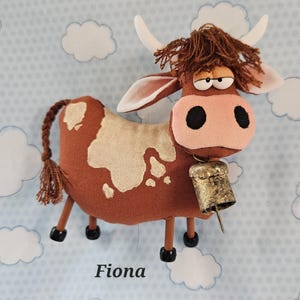 May include: A handmade brown cow plush toy with white spots, a pink nose, and a bell. The cow has a whimsical design with a fluffy brown mane, white horns, and the name "Fiona" printed below.