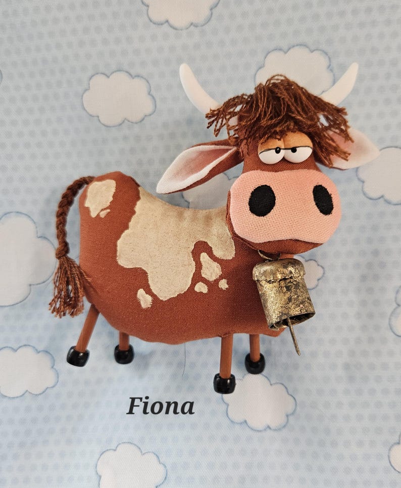 May include: A handmade brown cow plush toy with white spots, a pink nose, and a bell. The cow has a whimsical design with a fluffy brown mane, white horns, and the name "Fiona" printed below.