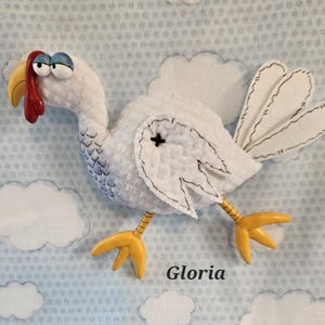 May include: A white turkey plushie with a red wattle, yellow beak and feet, and blue eyes. The turkey has a black stitched outline and a black cross on its wing. The background is a light blue fabric with white cloud shapes and the name "Gloria".