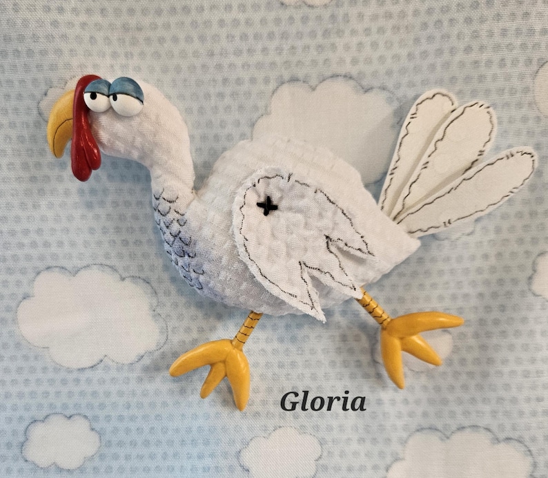 May include: A white turkey plushie with a red wattle, yellow beak and feet, and blue eyes. The turkey has a black stitched outline and a black cross on its wing. The background is a light blue fabric with white cloud shapes and the name "Gloria".