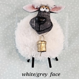 May include: A plush sheep figurine with a white, fluffy body and a gray and white patterned face. It has black legs, pink-lined ears, and a gold-colored bell around its neck. The text "white/grey face" is at the bottom.