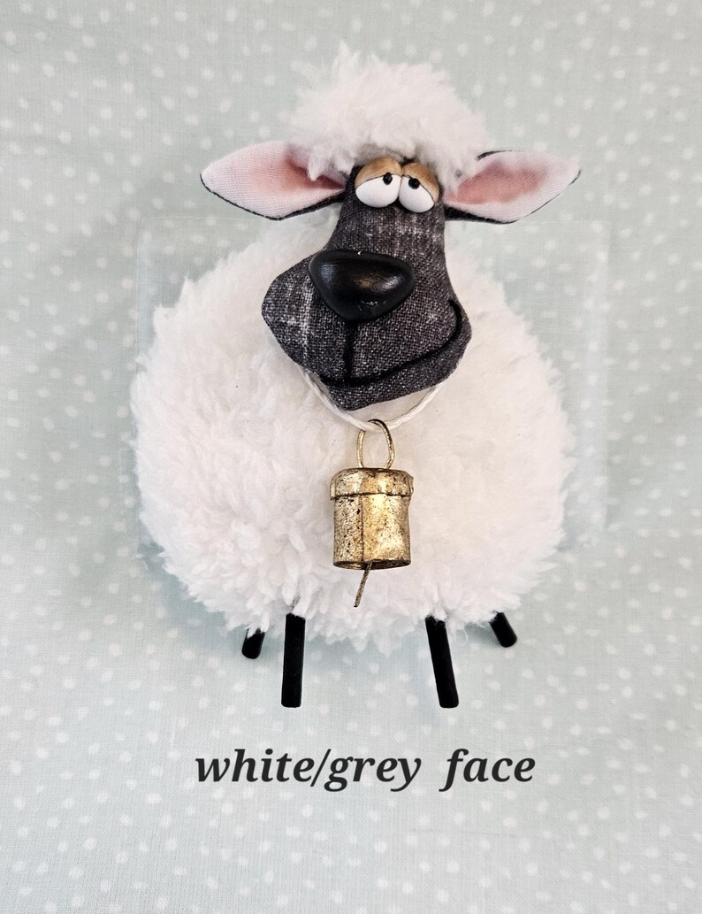 May include: A plush sheep figurine with a white, fluffy body and a gray and white patterned face. It has black legs, pink-lined ears, and a gold-colored bell around its neck. The text "white/grey face" is at the bottom.