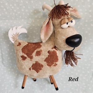 May include: A whimsical stuffed goat with a tan body and brown spots. It has a black nose, large googly eyes, and a brown fringe beard. The goat stands on four wooden legs and has a white tail. The word "Red" is written on the bottom right.