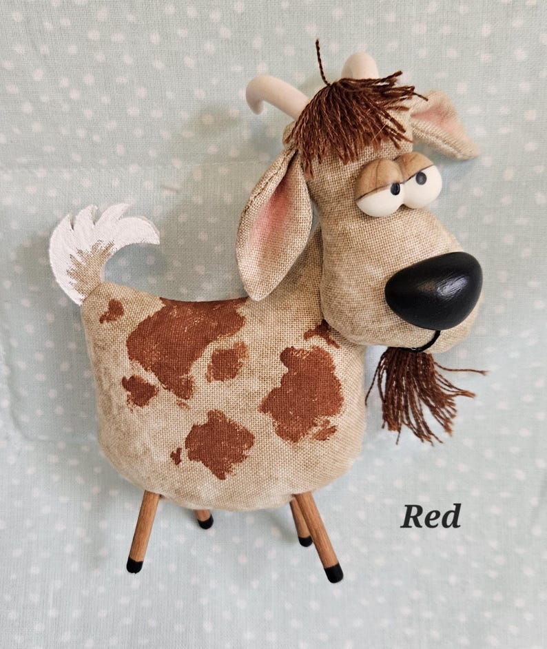 May include: A whimsical stuffed goat with a tan body and brown spots. It has a black nose, large googly eyes, and a brown fringe beard. The goat stands on four wooden legs and has a white tail. The word "Red" is written on the bottom right.