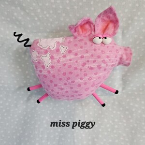 May include: A pink stuffed pig with large ears, googly eyes, and a curly tail. The pig has pink polka dots and white patches. The text "miss piggy" is printed below the pig.