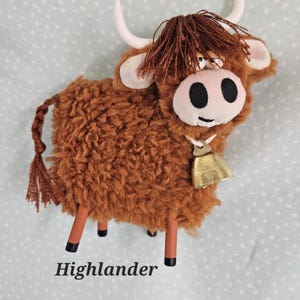 May include: A brown plush Highland cow with white horns, a bell, and the word "Highlander" at the bottom. The cow has a fluffy body, a brown fringe, and black eyes. The cow is standing on four brown legs.