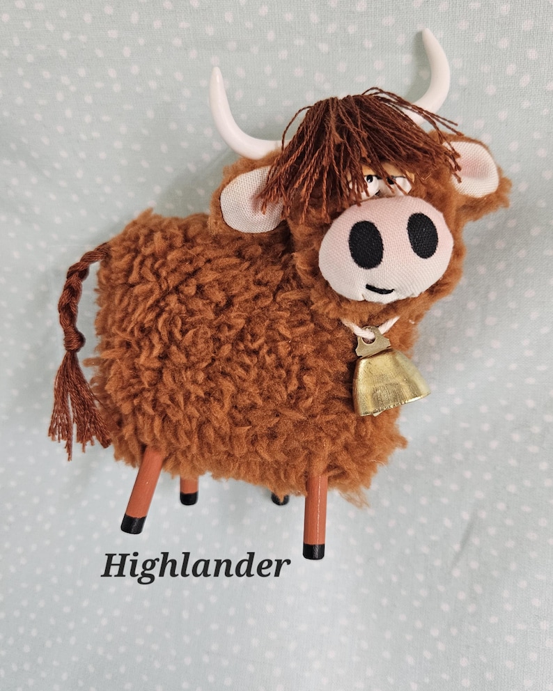 May include: A brown plush Highland cow with white horns, a bell, and the word "Highlander" at the bottom. The cow has a fluffy body, a brown fringe, and black eyes. The cow is standing on four brown legs.