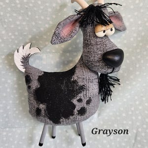 May include: A handmade goat figurine with a gray and black patterned body, black fringe, and a black nose. The goat has a white tail and horns, and is standing on gray legs. The name "Grayson" is printed on the bottom right.