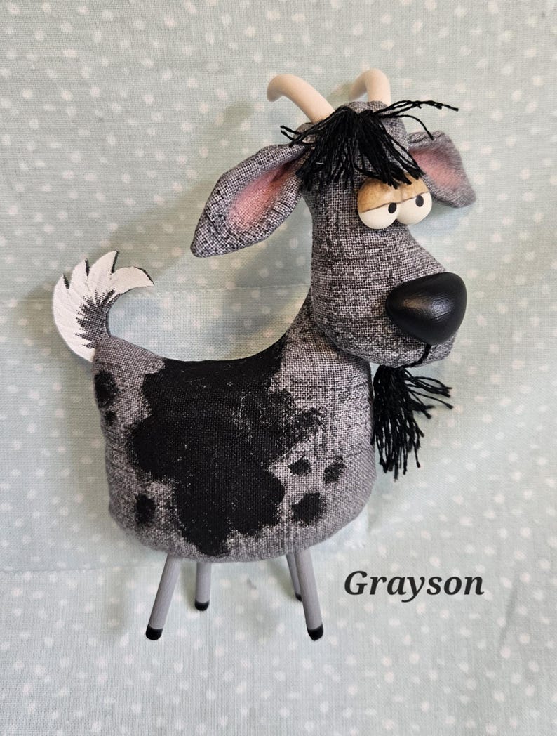 May include: A handmade goat figurine with a gray and black patterned body, black fringe, and a black nose. The goat has a white tail and horns, and is standing on gray legs. The name "Grayson" is printed on the bottom right.