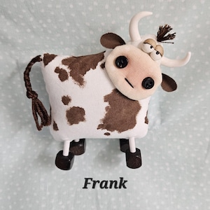 May include: A handmade stuffed cow toy with a white body and brown spots. It has a fabric face with button eyes and a stitched smile. The cow has white horns, brown ears, and a brown tail. The name "Frank" is printed below.
