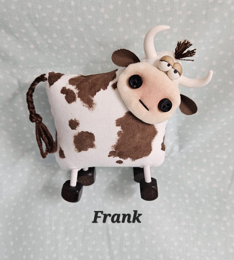 May include: A handmade stuffed cow toy with a white body and brown spots. It has a fabric face with button eyes and a stitched smile. The cow has white horns, brown ears, and a brown tail. The name "Frank" is printed below.