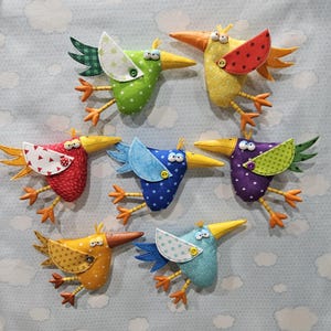 May include: Seven handcrafted fabric bird ornaments in various colors including green, yellow, red, blue, and purple. Each bird features a patterned wing attached with a button, a long beak, two googly eyes, and orange fabric feet.