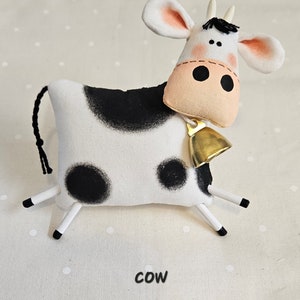 May include: A handmade stuffed cow toy with a black and white pattern. The cow has a pink nose, large black eyes, and a gold bell around its neck. The word "cow" is printed in black at the bottom.