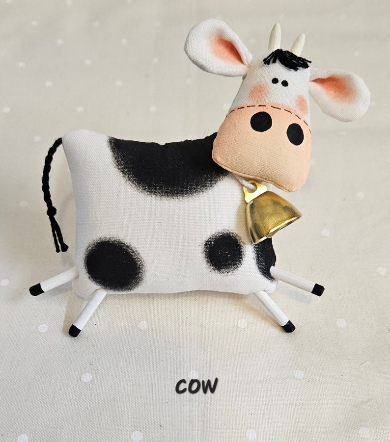 May include: A handmade stuffed cow toy with a black and white pattern. The cow has a pink nose, large black eyes, and a gold bell around its neck. The word "cow" is printed in black at the bottom.