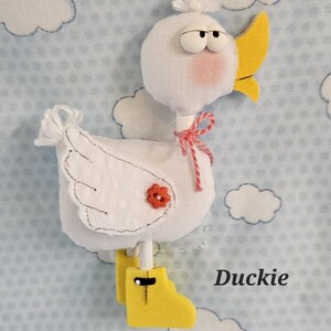 May include: A handmade white duck plushie with a yellow beak and boots, a red flower, and a pink bow. The duck has a fluffy white head and is set against a light blue background with white cloud shapes. The word "Duckie" is written in black.