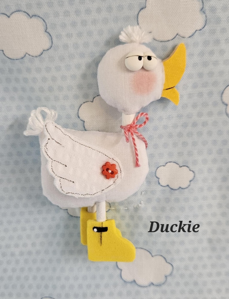 May include: A handmade white duck plushie with a yellow beak and boots, a red flower, and a pink bow. The duck has a fluffy white head and is set against a light blue background with white cloud shapes. The word "Duckie" is written in black.