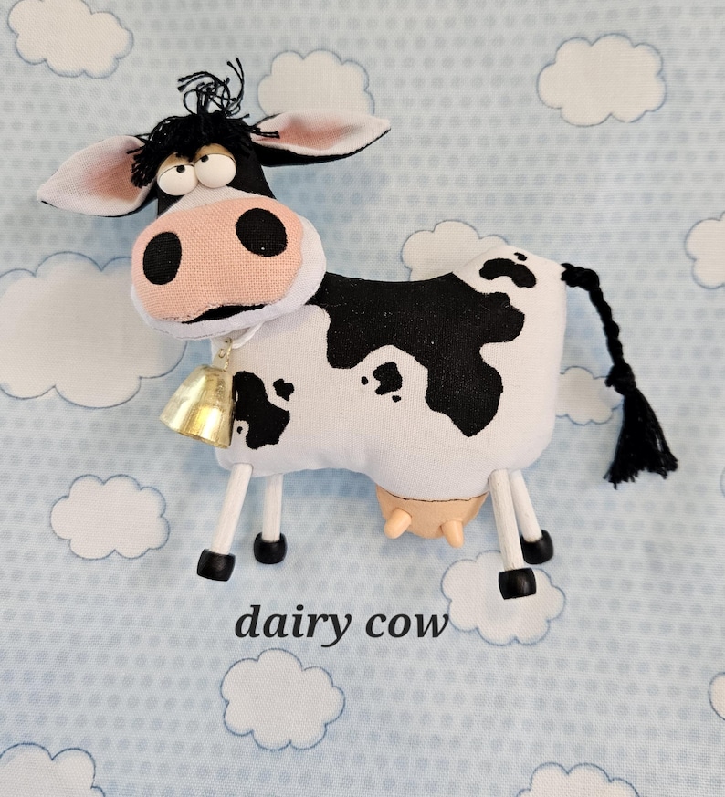 May include: A stuffed dairy cow with a bell around its neck. The cow is white with black spots and a pink nose. The text "dairy cow" is below the cow.
